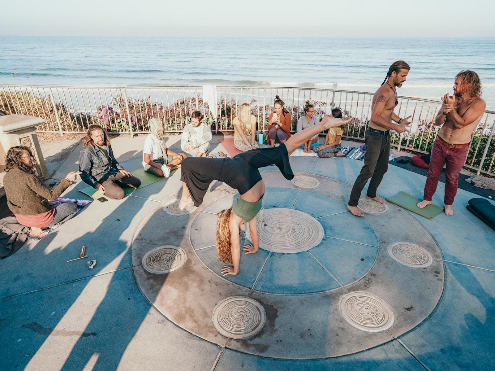 A small group on an oceanfront deck, one person upside down in a handstand while the others cheer